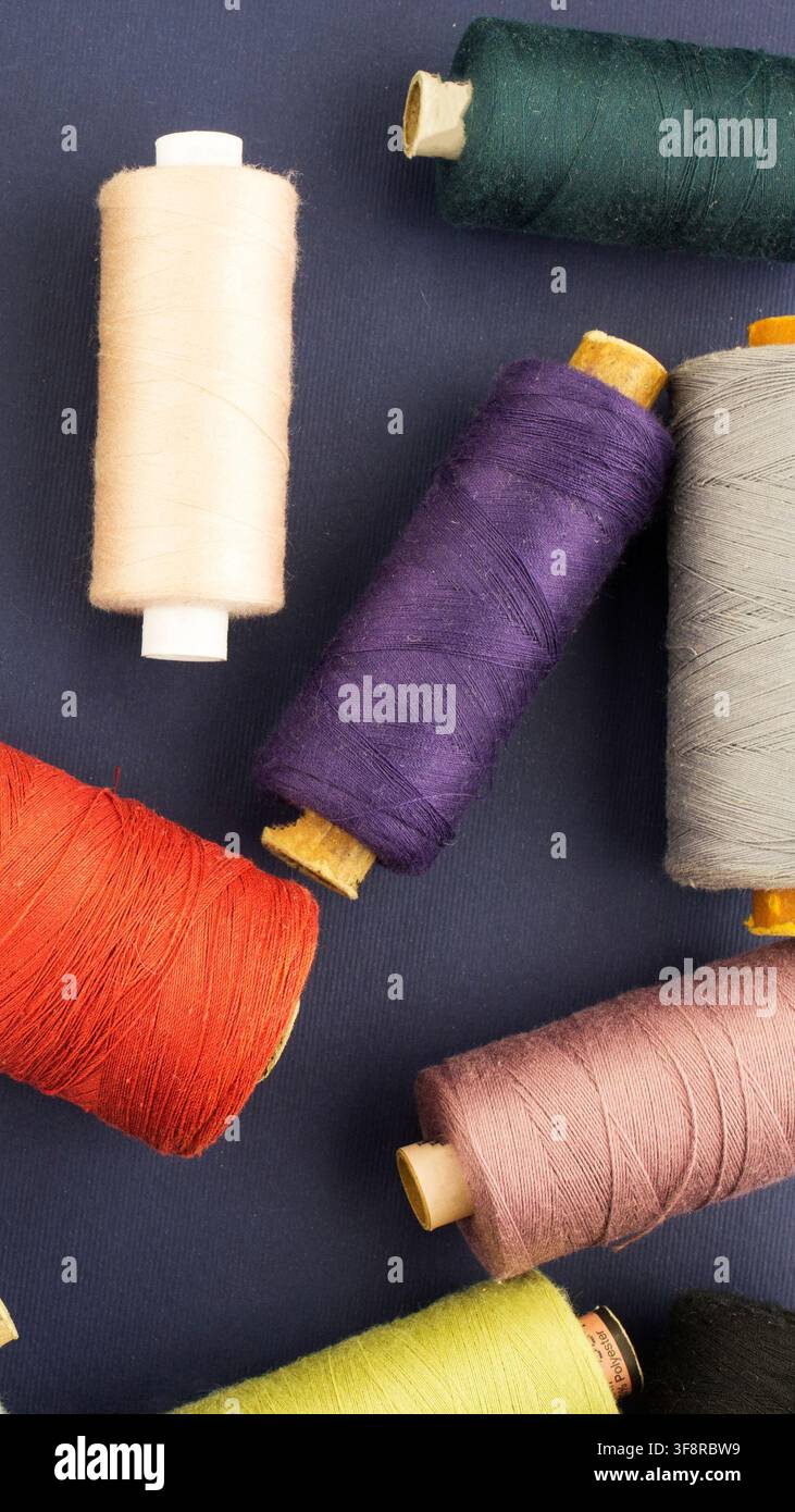 Variété de bobines de fil dans différentes teintes vives et pastel, notamment beige, orange, violet, vert et rose, étalées sur un bleu foncé texturé Banque D'Images