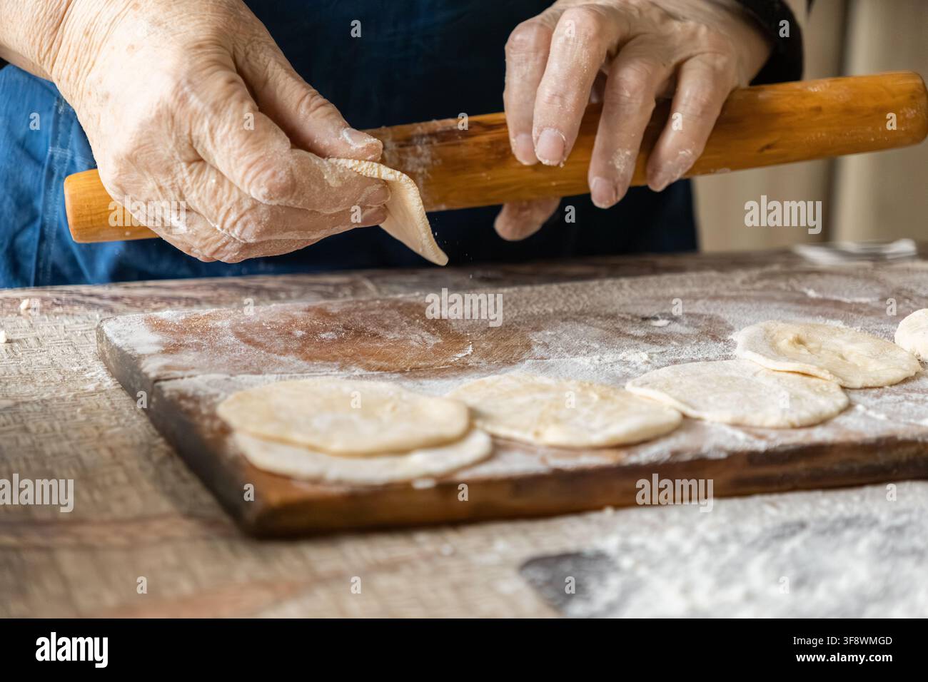 Des mains âgées pressent la pâte avec un rouleau en bois en préparant des boulettes maison sur une planche à découper farinée, mettant en valeur des techniques de cuisine authentiques et un Banque D'Images