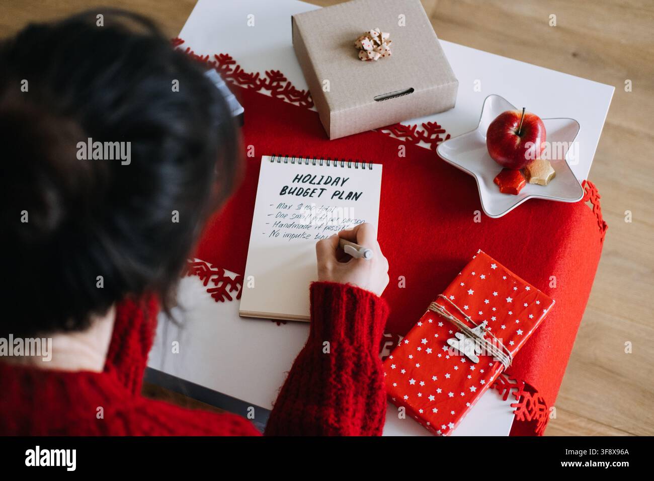 Femme écrit dans un carnet avec HOLIDAY BUDGET PLAN sur la table à côté de cadeaux et d'un panneau COZY CHRISTMAS. Économiser de l'argent pour Noël, fixation d'objectifs financiers Banque D'Images