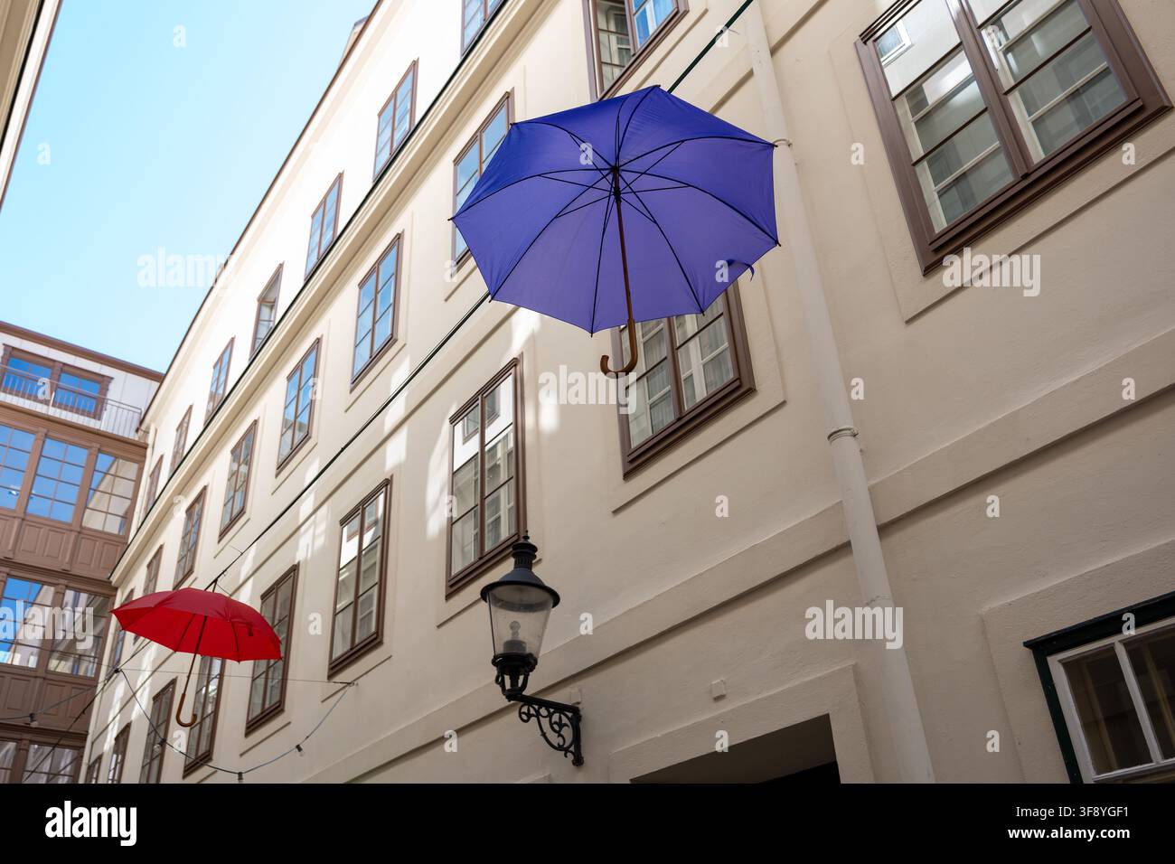 Un parapluie bleu et un parapluie rouge sont suspendus au-dessus d'une cour étroite entre des bâtiments aux couleurs claires sous un ciel dégagé. Banque D'Images