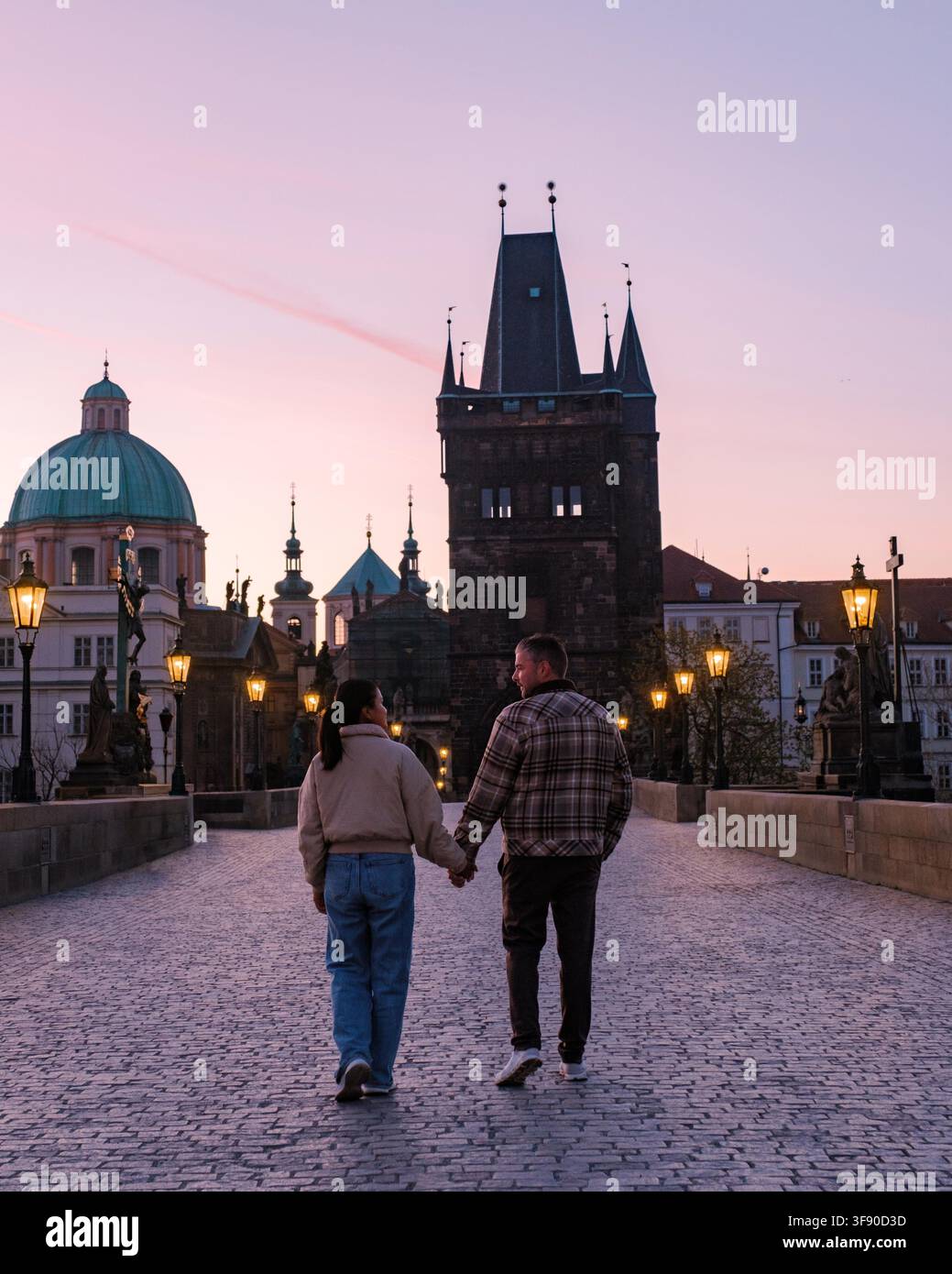 Un couple profite d'une promenade romantique en soirée sur le pont Charles à Prague. Le coucher de soleil projette une lueur chaude sur le paysage urbain, accentuant le charme de ce lieu historique rempli de touristes Banque D'Images
