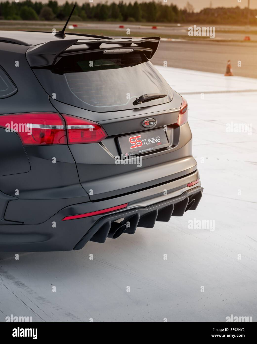 Arrière d'une Ford Focus break noire satinée modifiée avec un grand aileron de toit et un diffuseur agressif ainsi que des embouts d'échappement doubles. Piste de course sinueuse en arrière-plan Banque D'Images