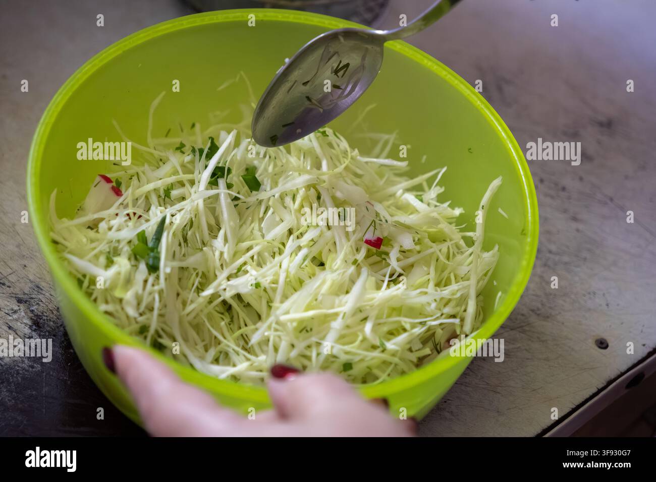 Une personne mélange du chou blanc tranché avec des herbes fraîches dans un bol en plastique vert citron pendant la cuisson, mettant en avant des habitudes alimentaires saines et des processus culinaires Banque D'Images