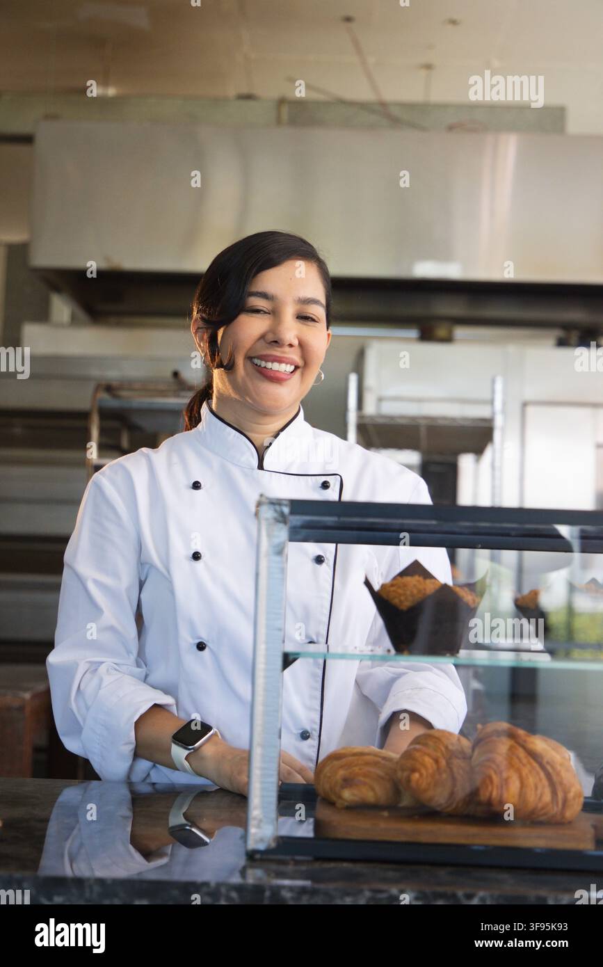 Chef d’âge moyen en blouse blanche et montre connectée derrière une vitrine dans une boulangerie proposant des croissants et des muffins Banque D'Images