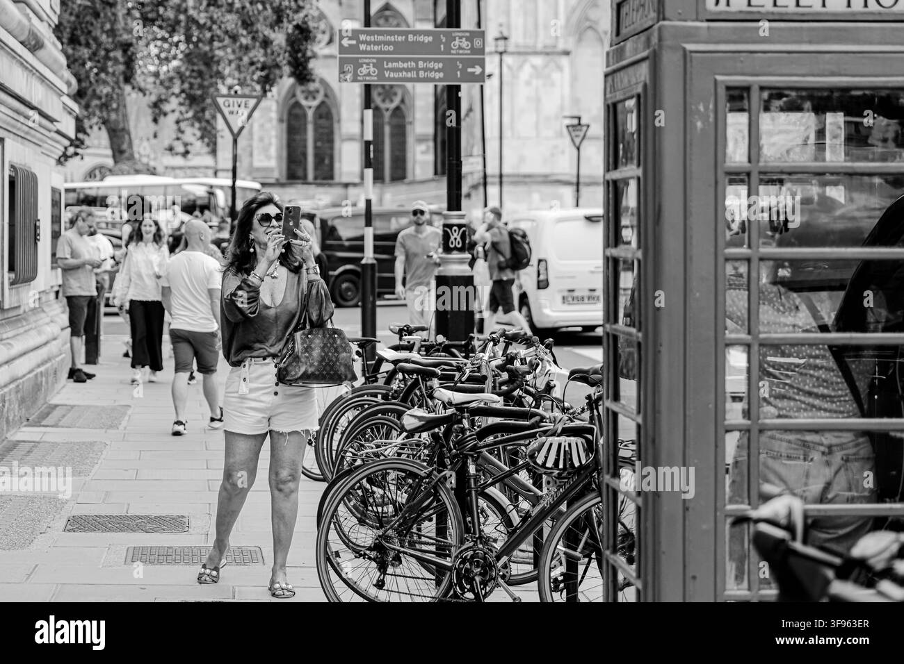 Une femme passe devant des vélos garés et un kiosque téléphonique traditionnel sur un trottoir animé du centre de Londres. Banque D'Images