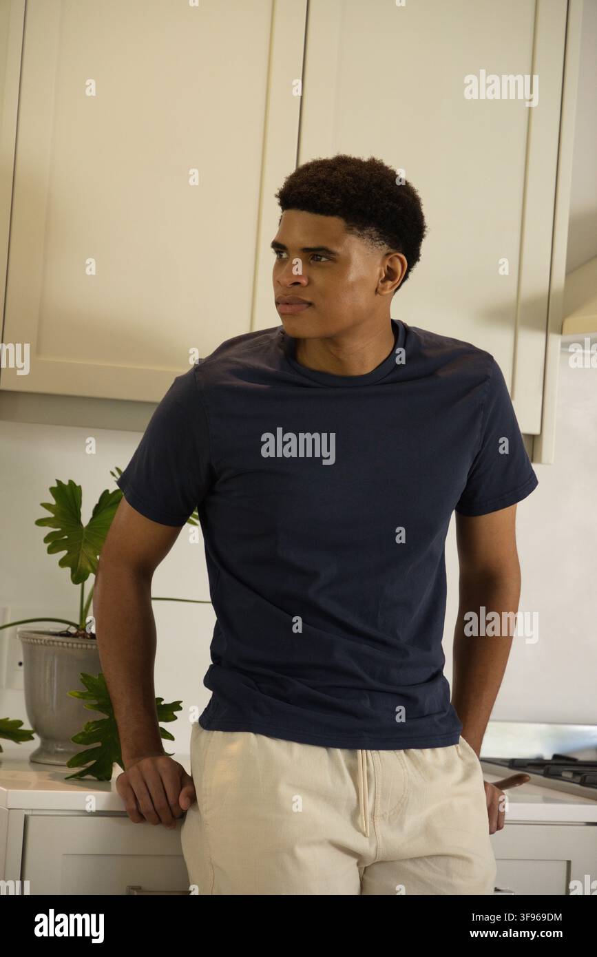 Homme afro-américain début de la vingtaine appuyé sur un comptoir dans la cuisine portant un T-shirt bleu marine, plante et poêle Banque D'Images