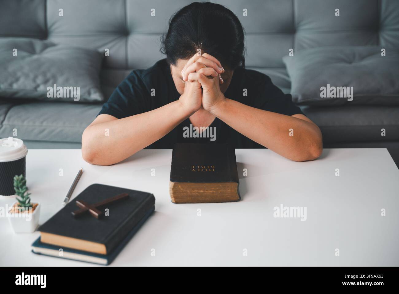 Femme chrétienne d'âge moyen priant la tête inclinée et les mains jointes à une table avec une Bible, des livres et une croix en bois, exprimant la foi, l'espoir, s Banque D'Images