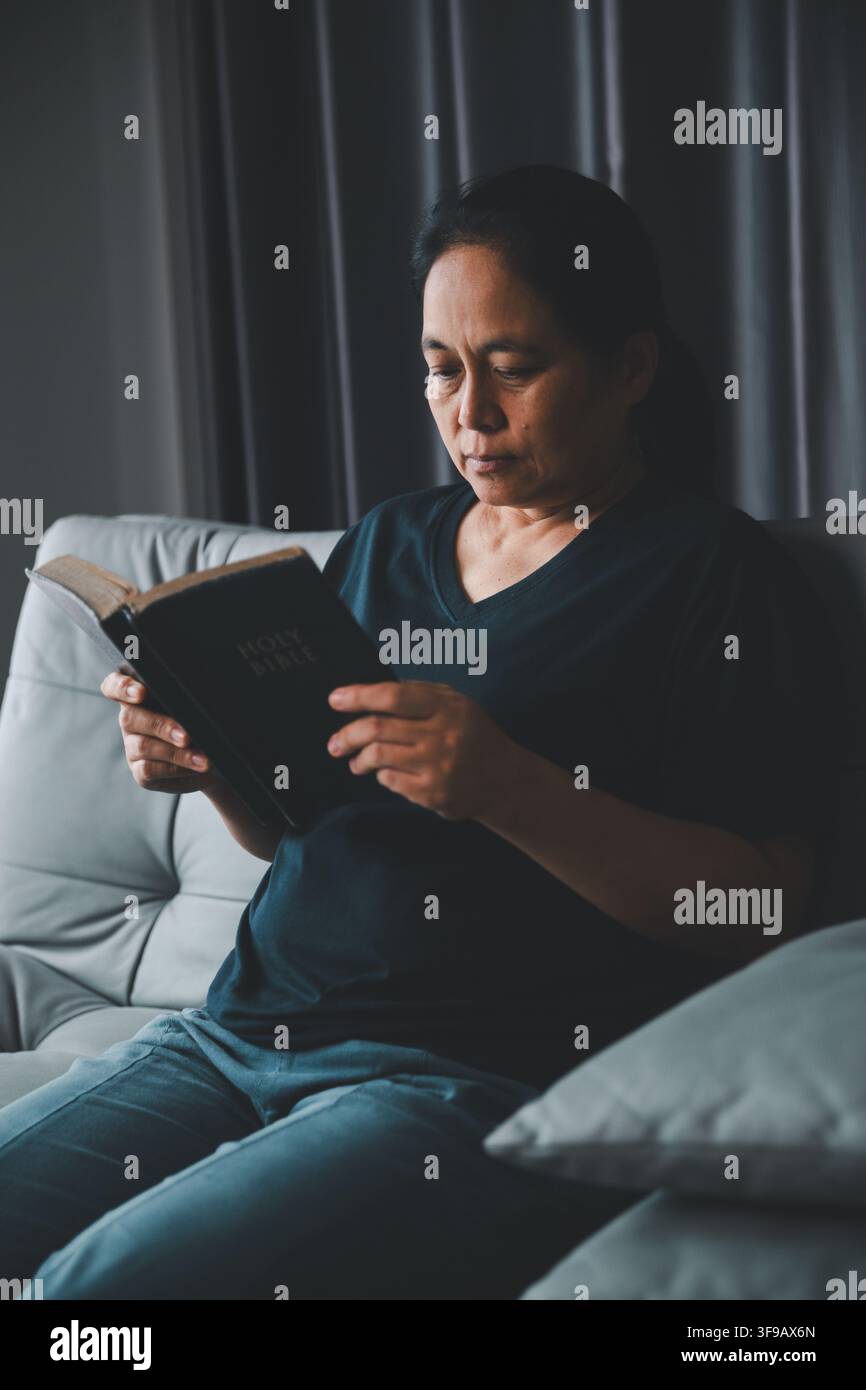Femme chrétienne d'âge moyen assise sur un canapé et lisant la Sainte Bible à la maison, représentant la foi personnelle, la dévotion, la réflexion spirituelle, le réconfort Banque D'Images