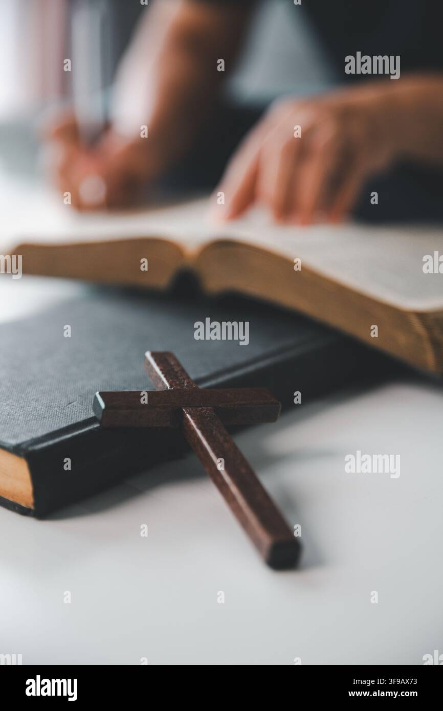 Croix chrétienne en bois à côté d'une Bible ouverte tandis qu'une personne écrit des notes d'étude en arrière-plan, représentant l'étude des Écritures, la dévotion, l'apprentissage Banque D'Images