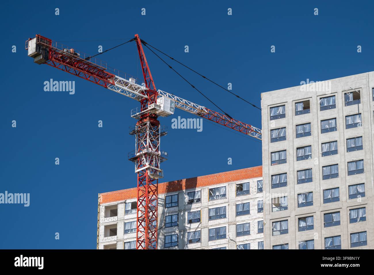 Dans une ville animée, une grue rouge vif se dresse fièrement contre un ciel bleu sans nuage, soutenant activement la construction d'un bâtiment moderne. Les ouvriers skient Banque D'Images