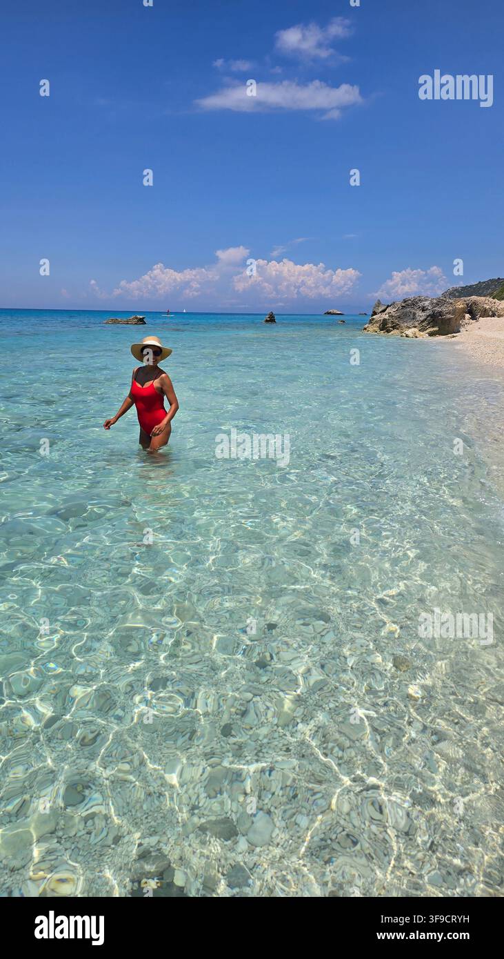 Une femme en maillot de bain rouge se promène dans les eaux cristallines de la plage de Gaidaros à Kalamitsi, Lefkada, Grèce. Le soleil brille de mille feux, illuminant le paysage côtier époustouflant. Banque D'Images