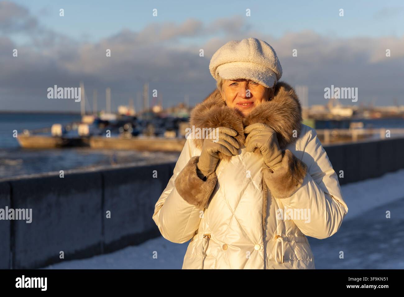 Belle femme mûre d'une beauté intemporelle regardant pensivement au loin sur un talus enneigé près du port maritime nordique pendant l'heure dorée. Banque D'Images