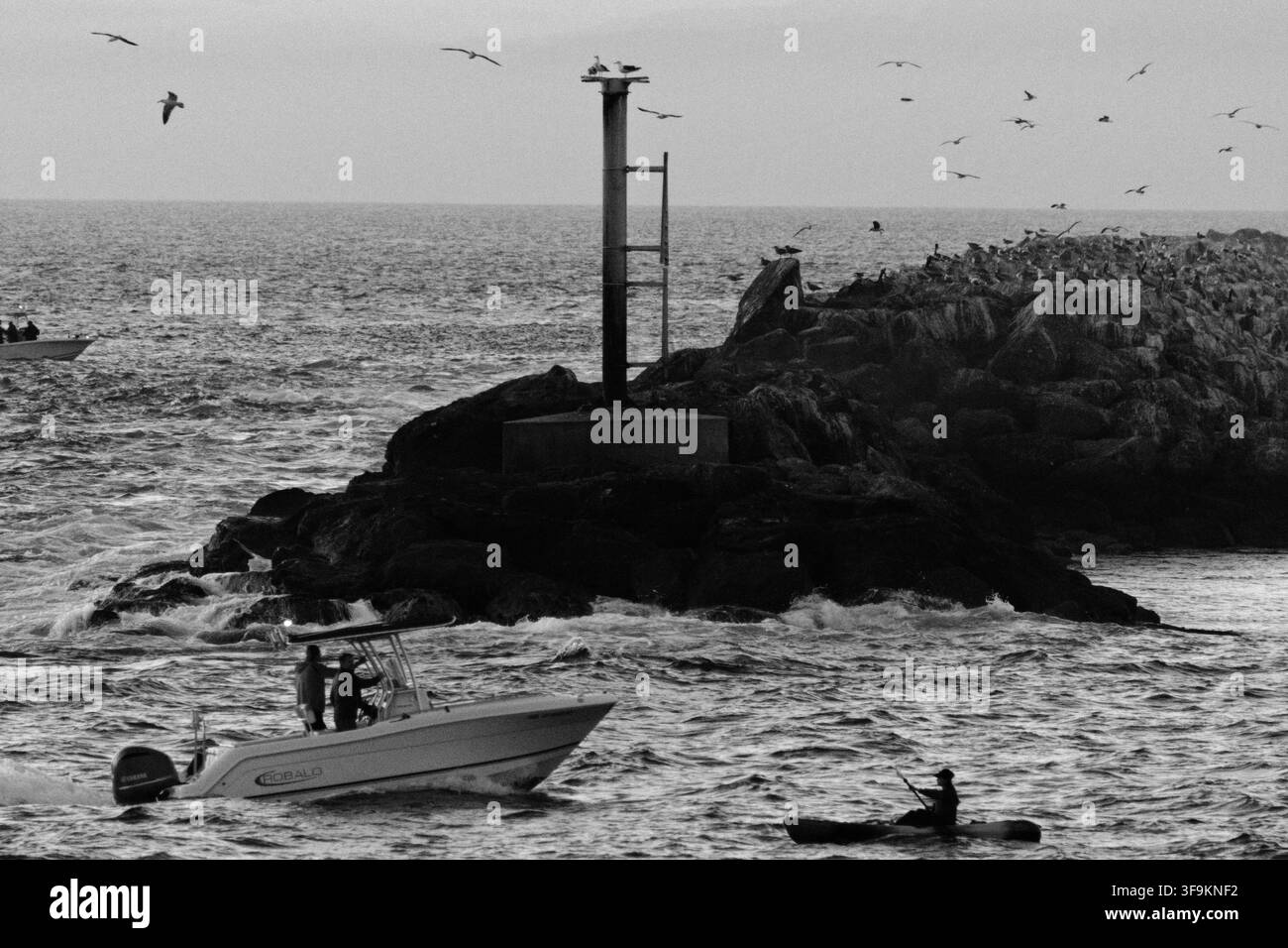 Bateau près d'une côte rocheuse avec phare. Les mouettes volent au-dessus d'une mer agitée. Deux personnes se tiennent sur un hors-bord, prêtes à explorer. Une autre personne flotte à proximité. L Banque D'Images