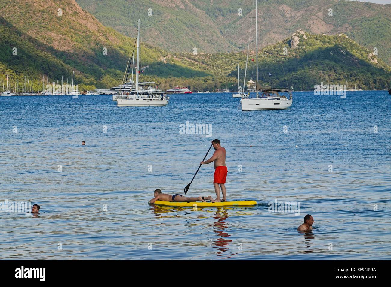 Jeune homme sur paddleboard dans la mer avec des yachts de luxe et des yachts ancrés devant des montagnes boisées vertes en arrière-plan à Marmaris, Turquie. Banque D'Images