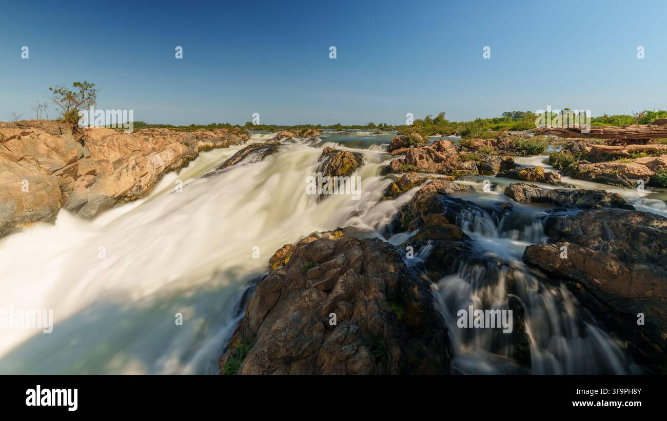 Vue en pose longue de larges cascades dévalant sur des rochers exposés aux chutes de Li Phi Somphamit, avec plusieurs chenaux sous un ciel bleu clair. Banque D'Images