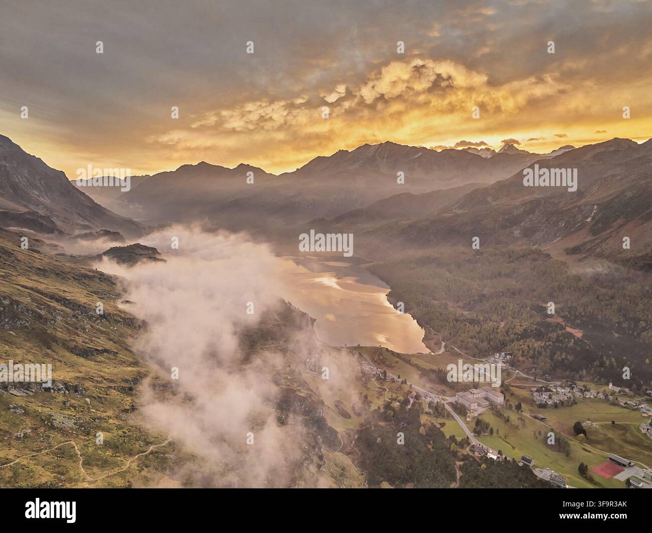 Vue aérienne de la vallée de l'Engadine avec un lac serein reflétant la lumière de l'heure dorée, entourée de montagnes couvertes de brume et de forêts luxuriantes dans les Grisons, S Banque D'Images