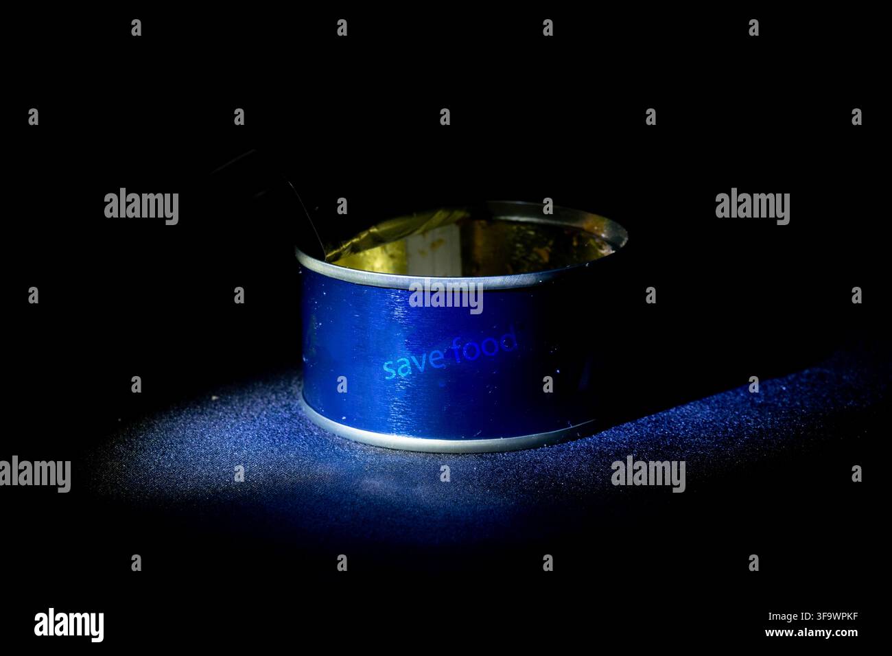 Boîte de conserve bleue ouverte avec un texte discret « Économiser les aliments » sous un projecteur directionnel sur fond sombre pour les campagnes de sécurité alimentaire et de conservation Banque D'Images