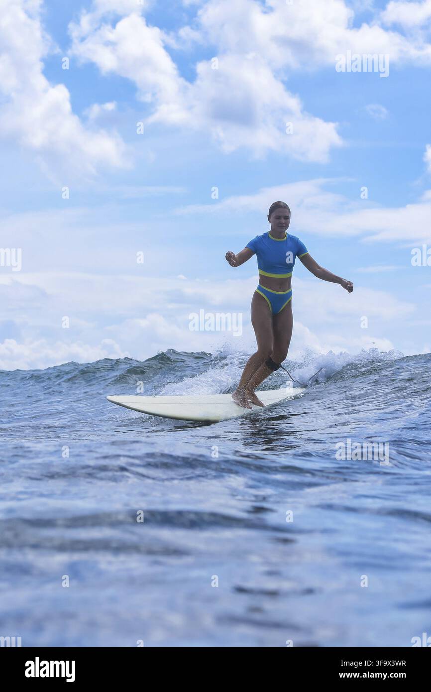 Surfeuse chevauchant une vague haute avec les bras levés, moment de triomphe culminant, ciel nuageux dramatiquement éclairé en contre-jour Banque D'Images