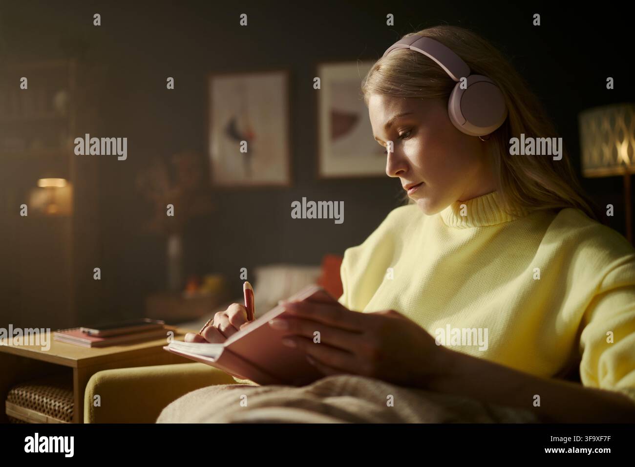 Jeune femme caucasienne portant des écouteurs et écrivant dans un carnet à la maison, étudiant ou tenant un journal. L'image représente le bien-être mental et l'apprentissage à distance, adaptée au marketing éducatif Banque D'Images