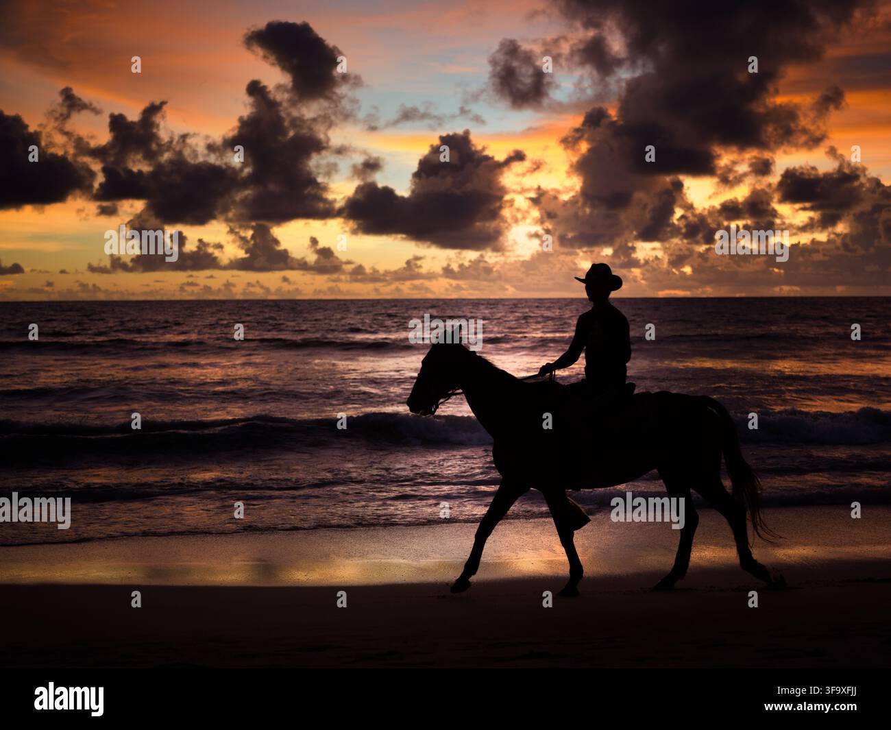 Cheval et cavalier avec chapeau de cowboy en silhouette devant un coucher de soleil rouge et orange vif sur une plage tropicale. Plage de Kamala, Phuket, Thaïlande. Banque D'Images