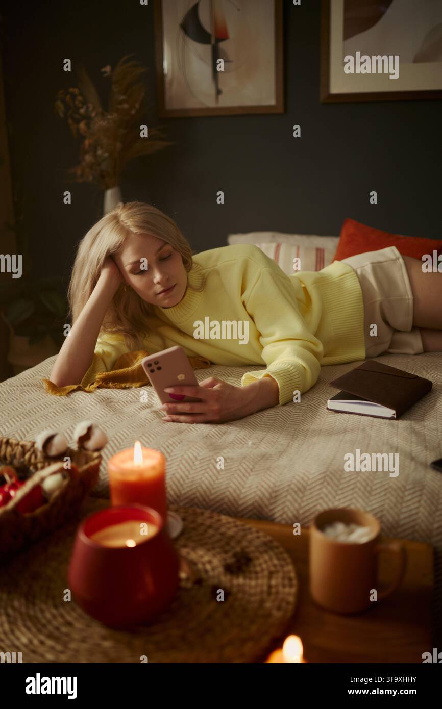 Image verticale d'une jeune femme caucasienne allongée sur un lit et utilisant un smartphone. L'image représente la vie numérique et la détente, adaptée au bien-être mental et au marketing de style de vie Banque D'Images