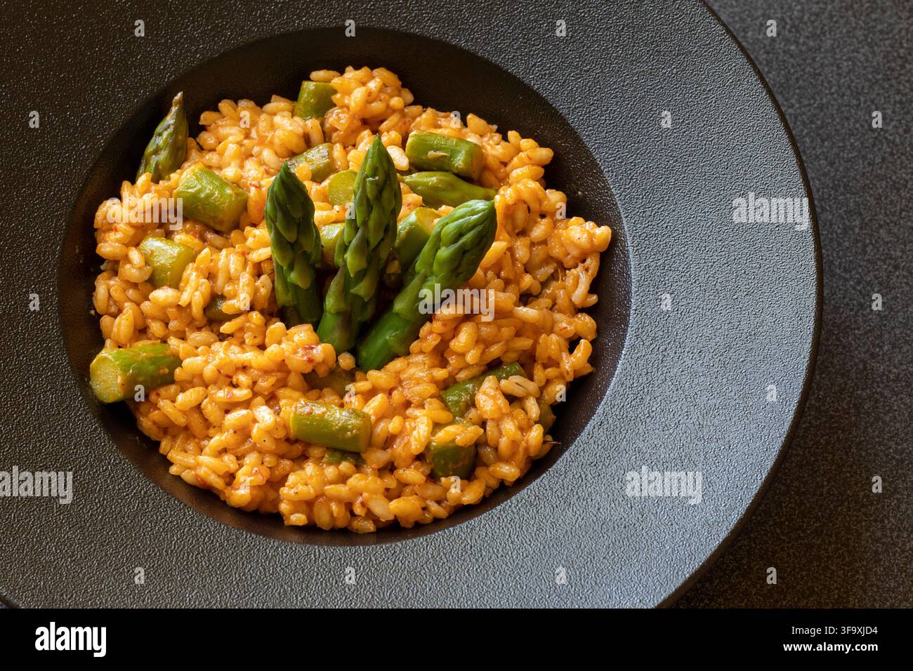 Vue aérienne d'un risotto crémeux à la nduja avec des pointes d'asperges vertes dans un bol à bord large noir mat posé sur un plan de travail sombre à pois Banque D'Images