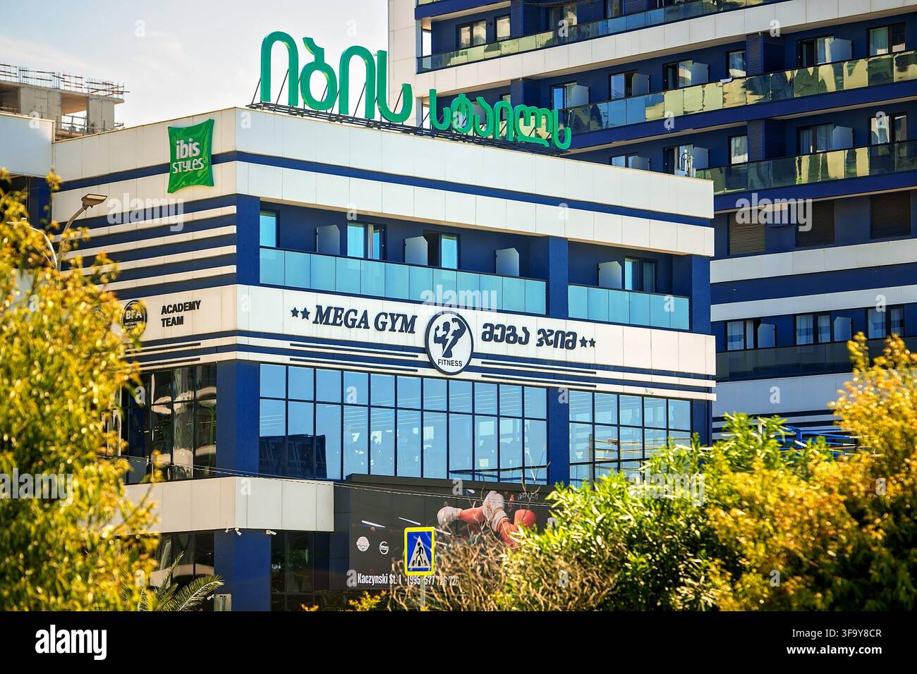 Batumi, Géorgie - 19 juillet 2026 : Bâtiment moderne avec enseigne Ibis Styles hôtel et club de fitness Mega Gym sur la façade vitrée encadrée par des arbres verts Banque D'Images