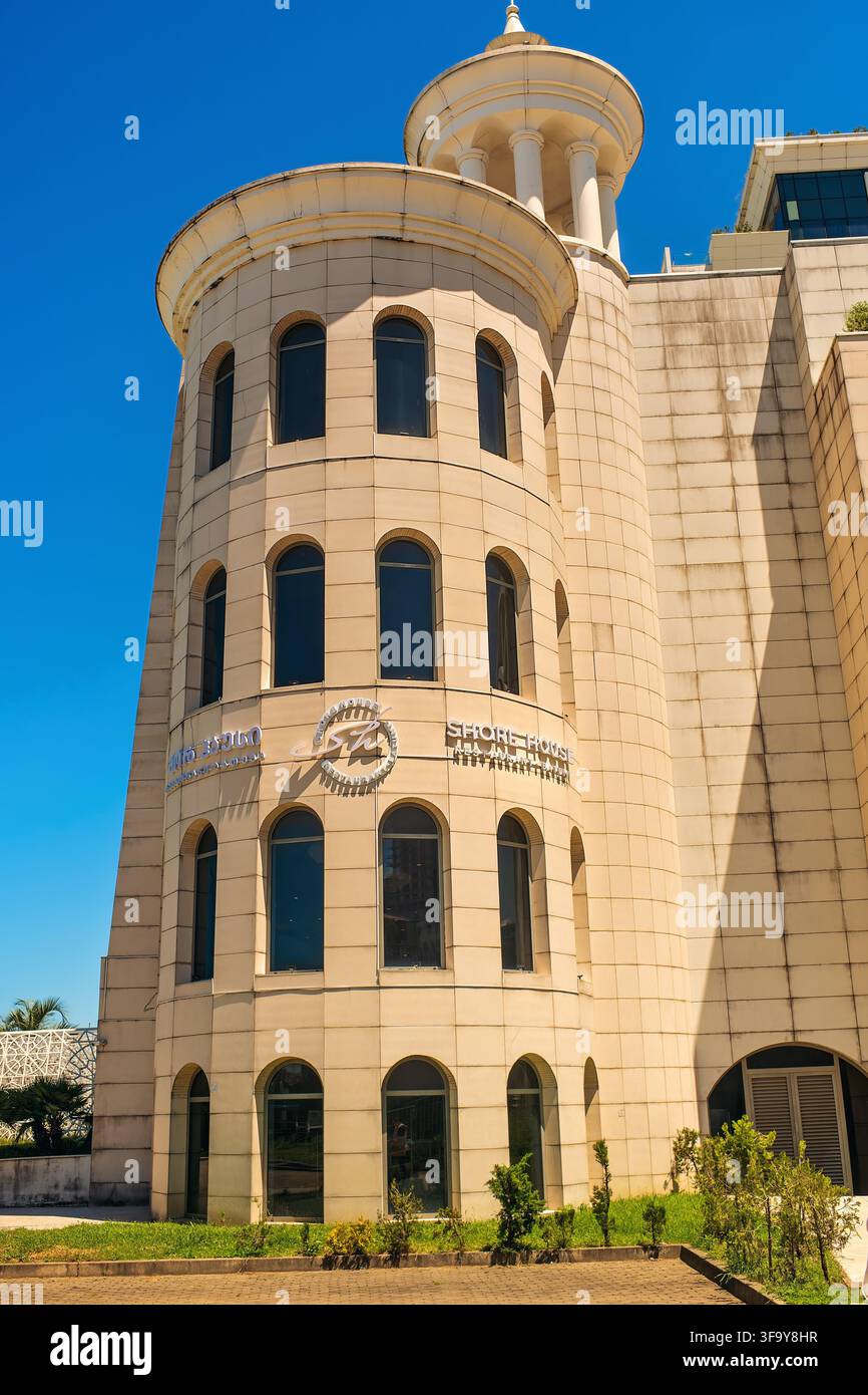 Batumi, Géorgie - 19 juillet 2026 : Façade d’un bâtiment moderne jaune avec une tour ronde, des fenêtres en arc et l’enseigne du restaurant Shore House sous un ciel bleu Banque D'Images