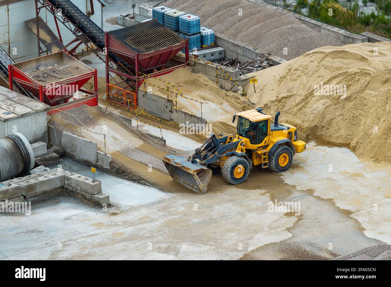 Vue aérienne d'un chargeur à roues jaune opérant dans une usine de ciment avec des tas de sable, de gravier et des équipements de convoyage industriels en Pologne. Banque D'Images