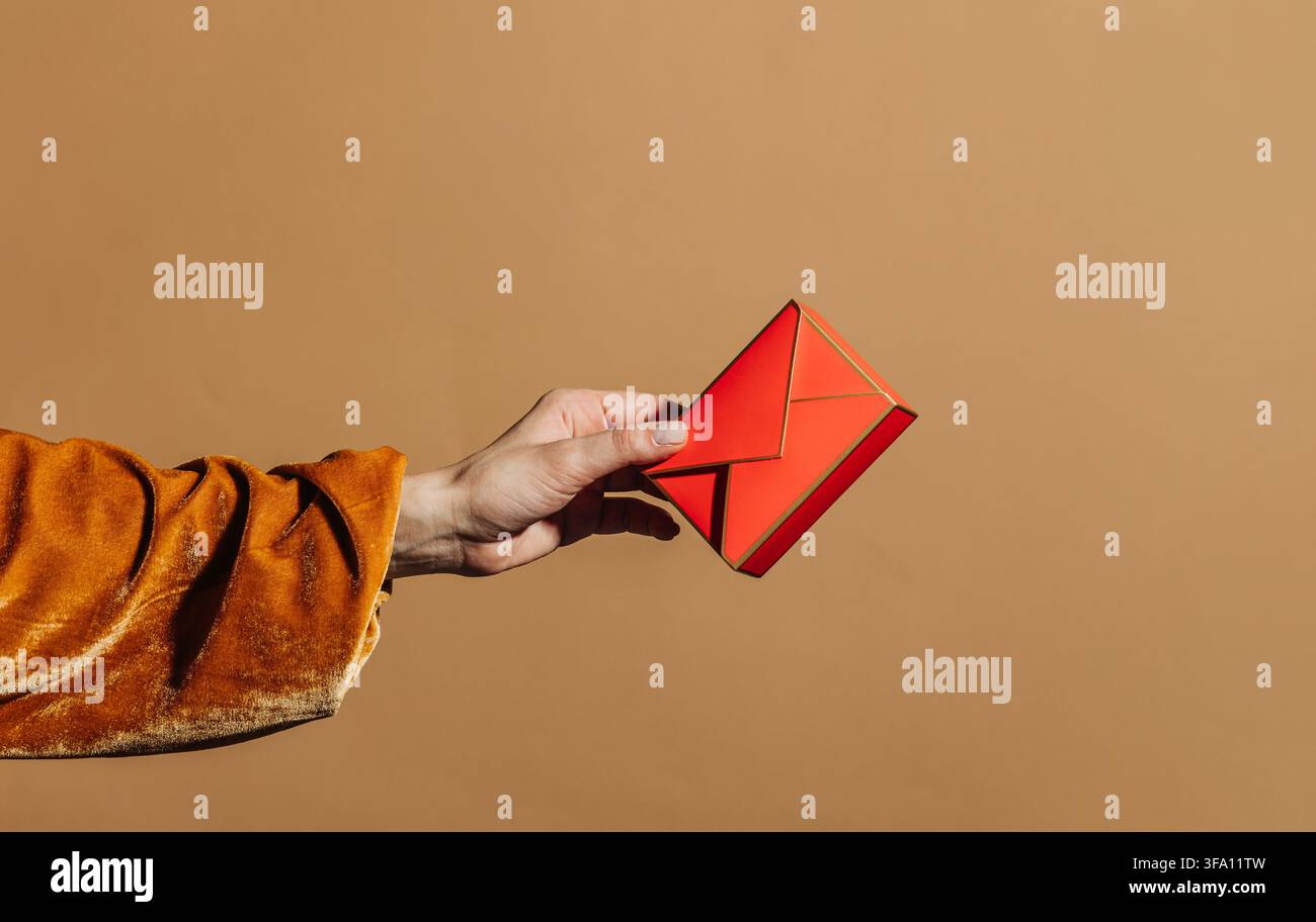 Close-up d'une main dans une manche en velours orange tenant un cadeau rouge géométrique en enveloppe contre un fond beige clair, avec un espace suffisant pour du texte de célébration Banque D'Images