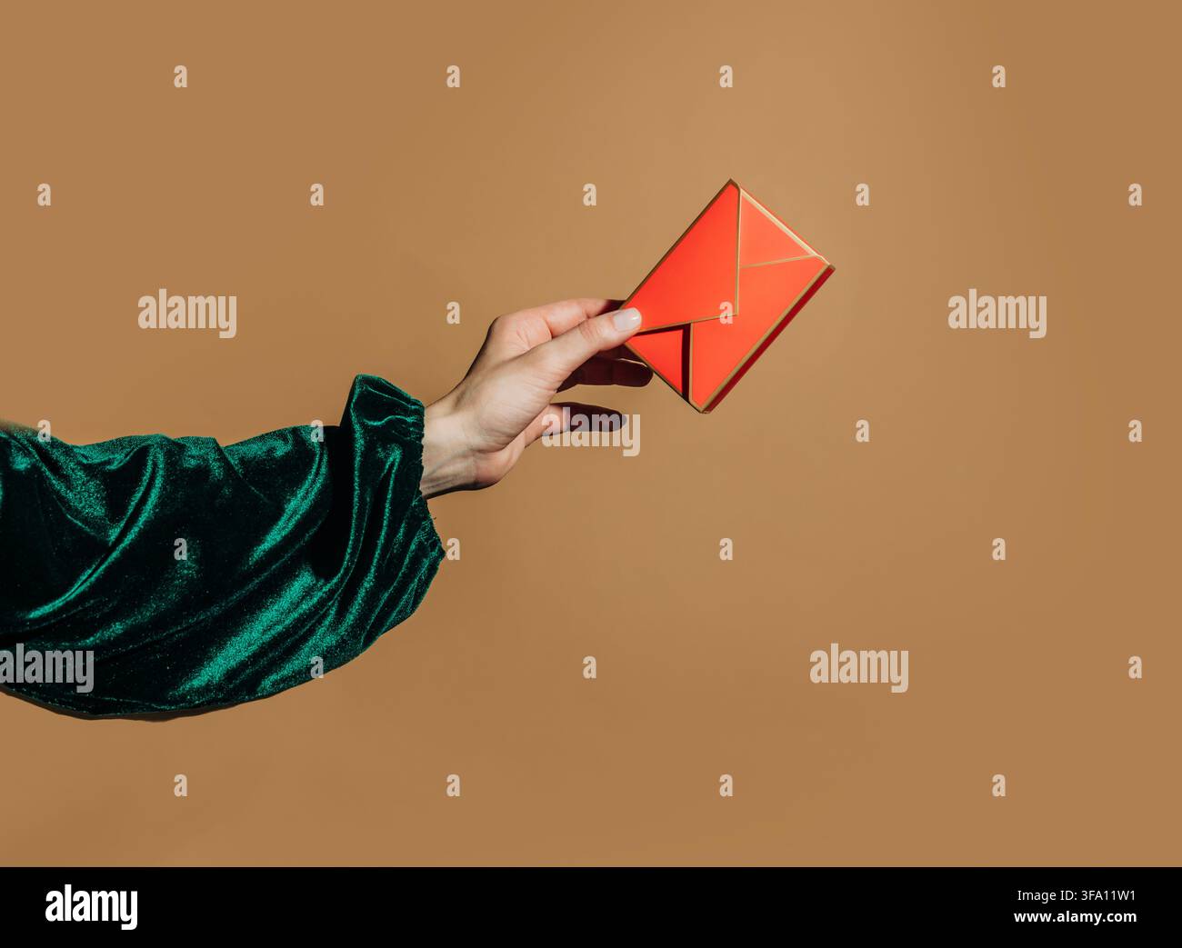Une main dans une manche en velours vert tient une enveloppe origami rouge vif contre un fond brun chaud, créant un concept festif pour les vœux, invitati Banque D'Images