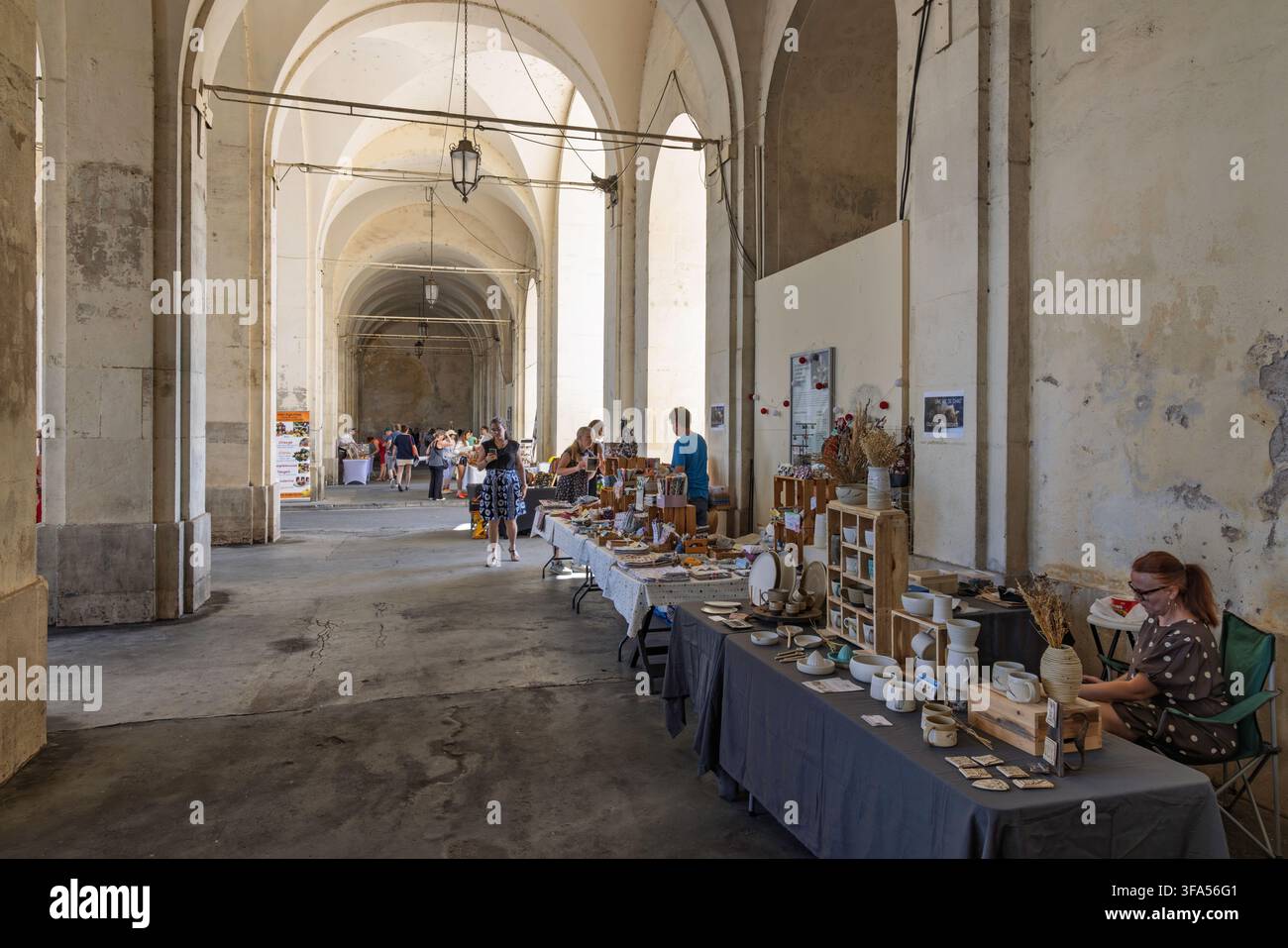 Nancy, France - 02 août 2026 : Étals de marché artisanal avec des céramiques et des produits locaux faits main sous des arcades en pierre historiques. Banque D'Images