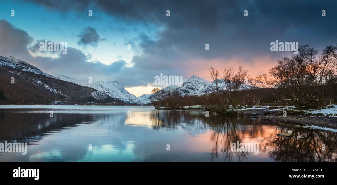 Un coucher de soleil panoramique hivernal éclaire des nuages épais au-dessus de montagnes enneigées se reflétant dans les eaux calmes du Llyn Padarn au Pays de Galles du Nord. Banque D'Images