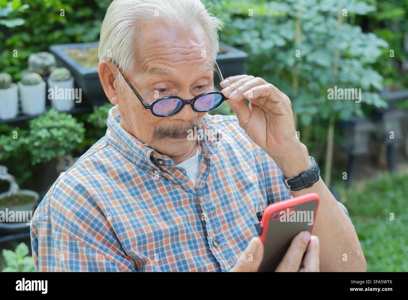 Homme asiatique âgé utilisant un smartphone, portant des lunettes et ayant une mauvaise vue en extérieur. Concept de problèmes de vue. Banque D'Images