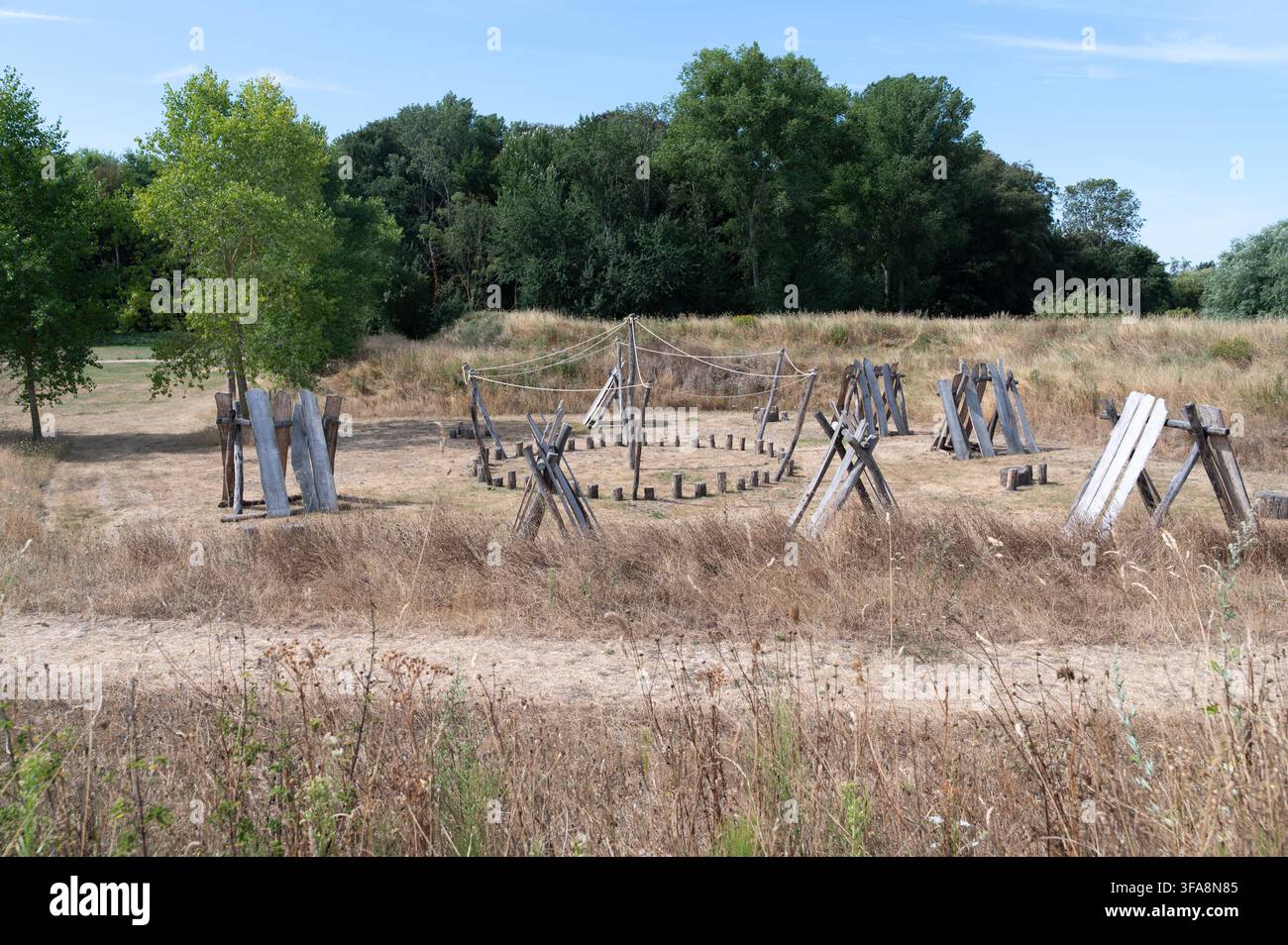 Une structure en bois extérieure située dans un champ herbeux sec avec des arbres clairsemés et un ciel bleu clair, présentant diverses formes géométriques disposées en un Banque D'Images