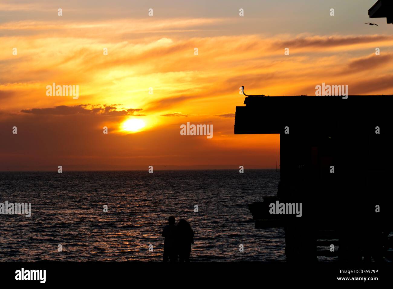 Le coucher de soleil peint l'océan dans des tons orange et dorés chauds. Silhouette d'un ponton et d'un couple sur le pont. Le couple se tient proche, profitant d'un moment paisible. Parfait pour ro Banque D'Images
