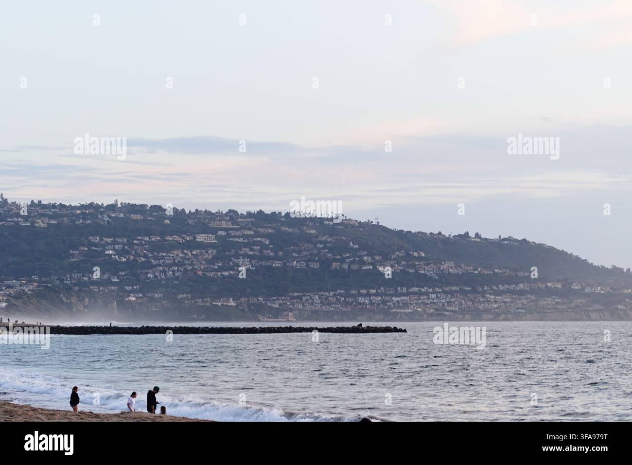 Scène côtière au crépuscule, mer calme rencontrant une rive sablonneuse. Ville des collines au loin, éclairage doux. Des gens se promènent près du bord de l'eau, détendus et paisibles. Idéal Banque D'Images