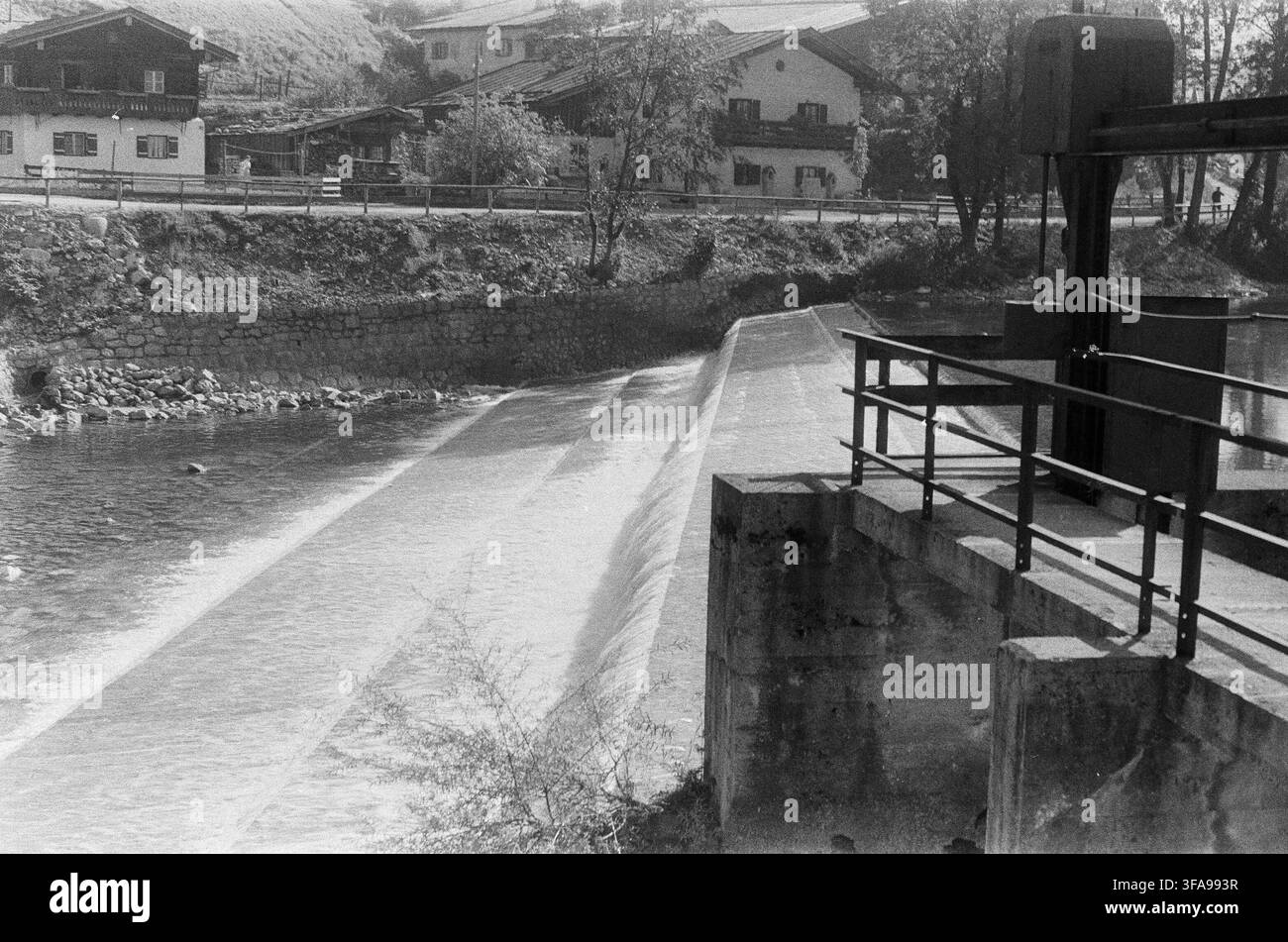 Un barrage en béton avec de l'eau qui coule dans un petit village alpin, avec des bâtiments traditionnels de style chalet et des murs de retenue en pierre. Classique du milieu du 20e siècle Banque D'Images
