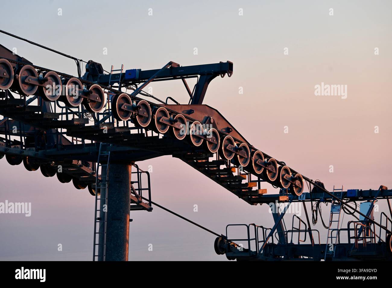 Photographie publicitaire en gros plan de machines industrielles lourdes pour un système de téléphérique ou de remonte-pente, devant un ciel doux au coucher du soleil. Le cliché met l'accent sur les mécanismes complexes, les supports en acier structurel et une longue ligne de roues métalliques rotatives et de câbles utilisés pour le transport. Banque D'Images