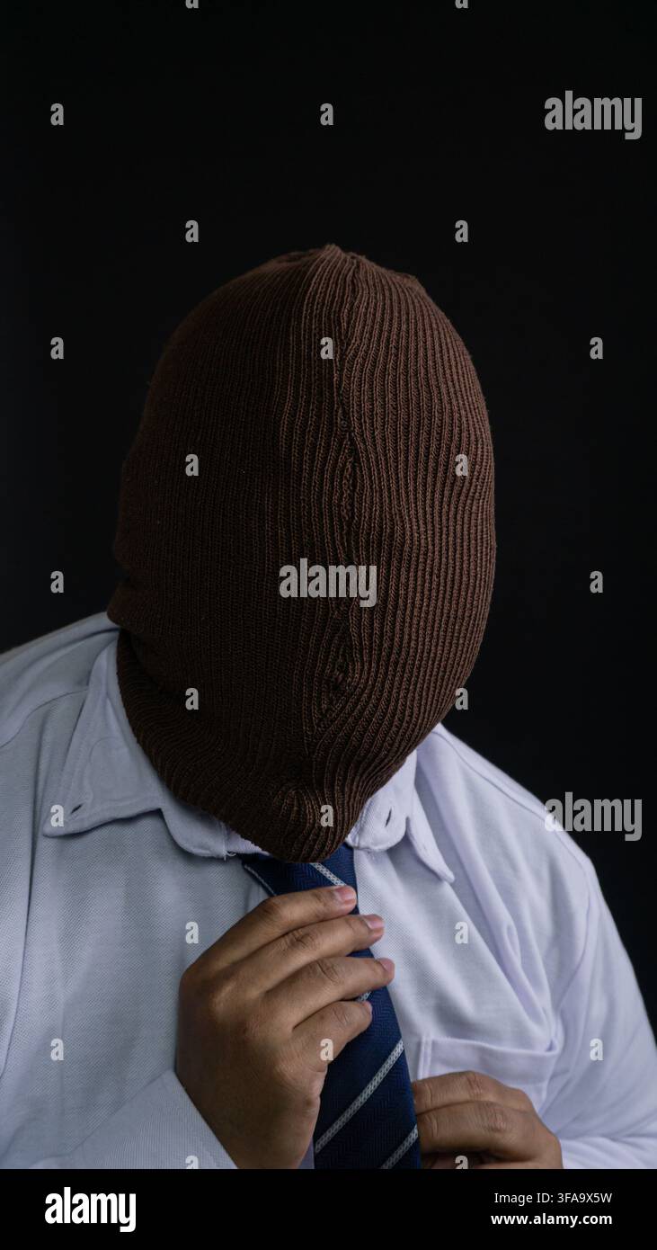 Un homme asiatique anonyme avec un visage entièrement dissimulé par une cagoule tricotée brune regarde vers le bas tout en ajustant soigneusement un n à rayures bleues Banque D'Images
