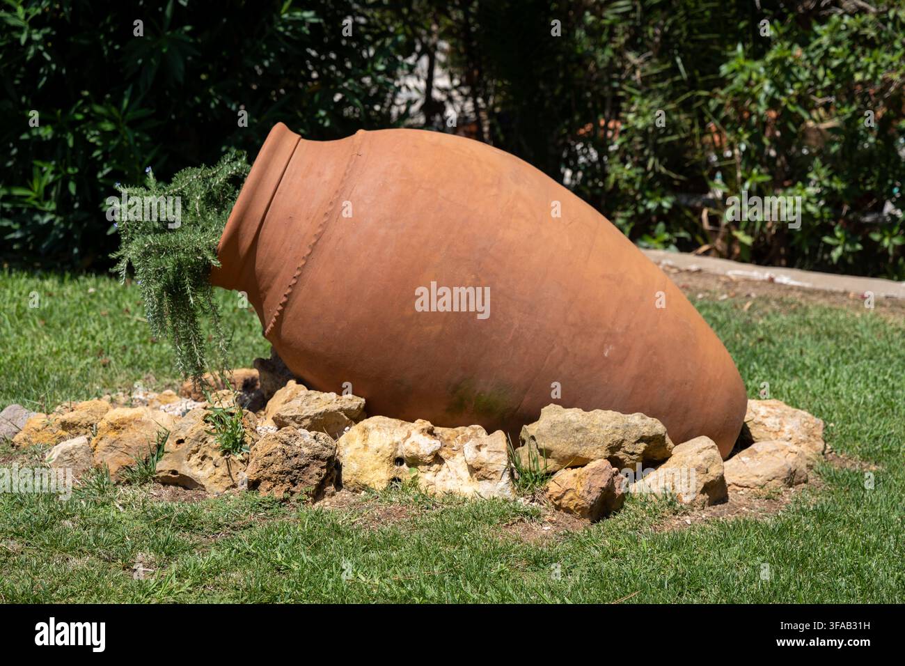 Un pot en terre cuite de style ancien gît sur le côté dans un jardin, entouré de rochers et d'herbe, avec une plante qui pousse à partir de son ouverture Banque D'Images