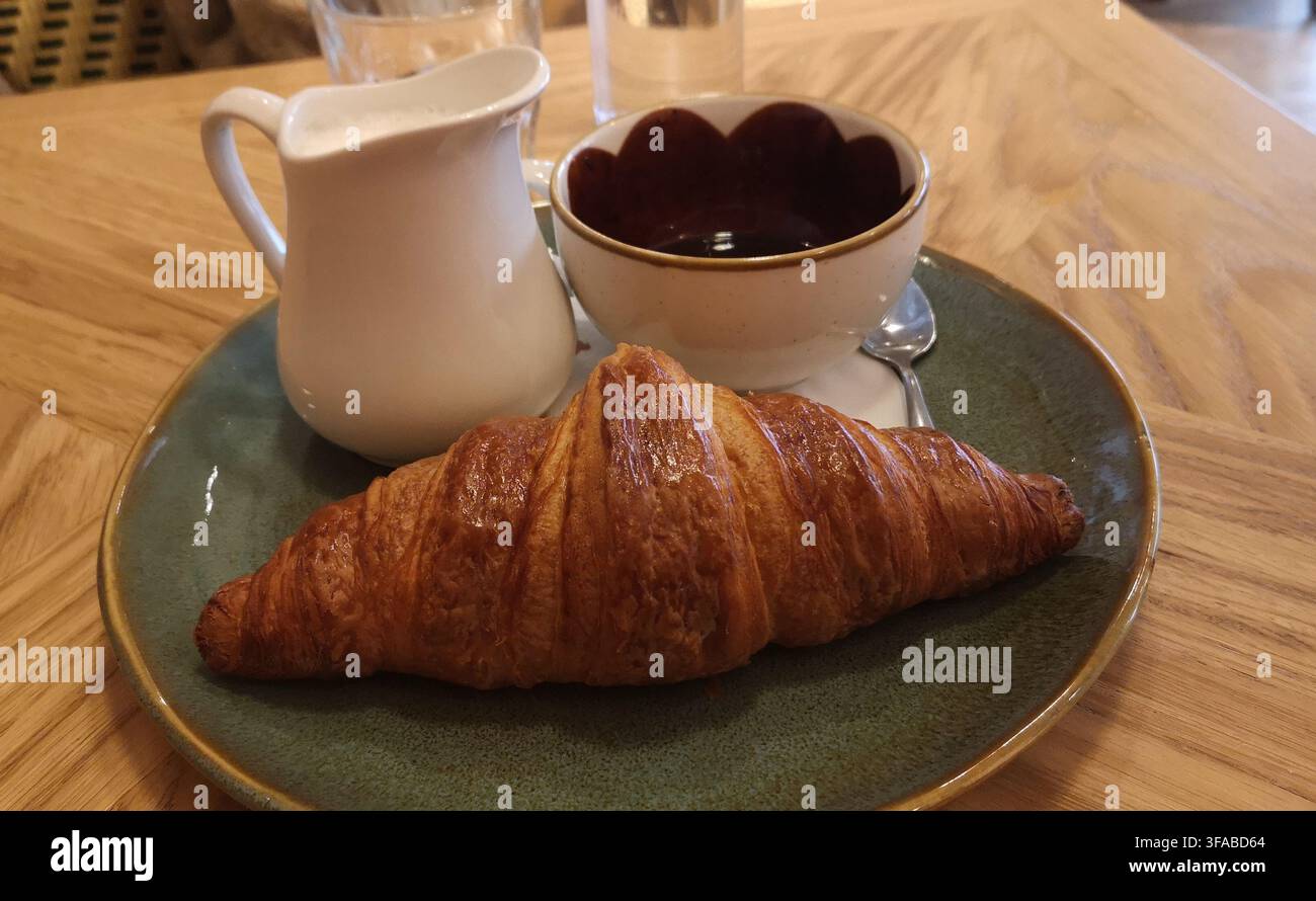 Petit-déjeuner continental européen confortable mettant en vedette un croissant feuilleté cuit au four, du lait chaud et un riche espresso sur une table en bois élégante. Banque D'Images