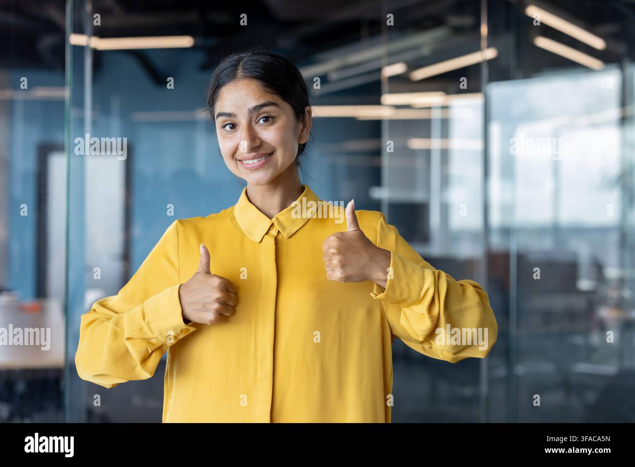 Portrait d'une jeune Indienne debout dans le bureau et montrant à la caméra un signe de réussite avec ses doigts. Banque D'Images