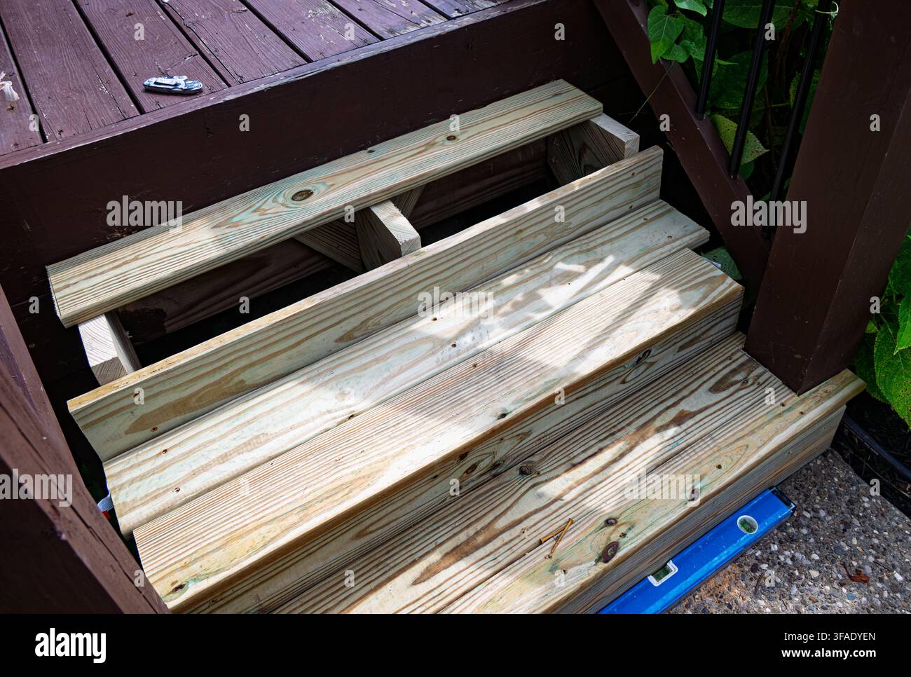 Remplacement d'escalier en bois en construction menant à une terrasse brun foncé. Une surface bleue repose sur le gravier à côté des marches. Une végétation verte encadre la sc Banque D'Images