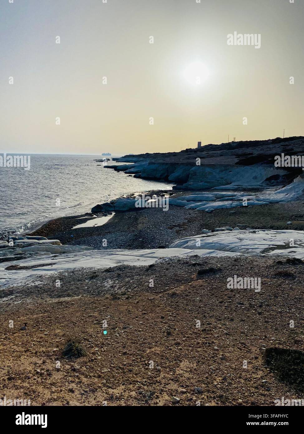 La lumière du soleil du soir tombe sur une rive rocheuse calme, où les rochers côtiers pâles rencontrent des galets sombres et une eau de mer douce. - Image de stock capturée avec un smartphone
