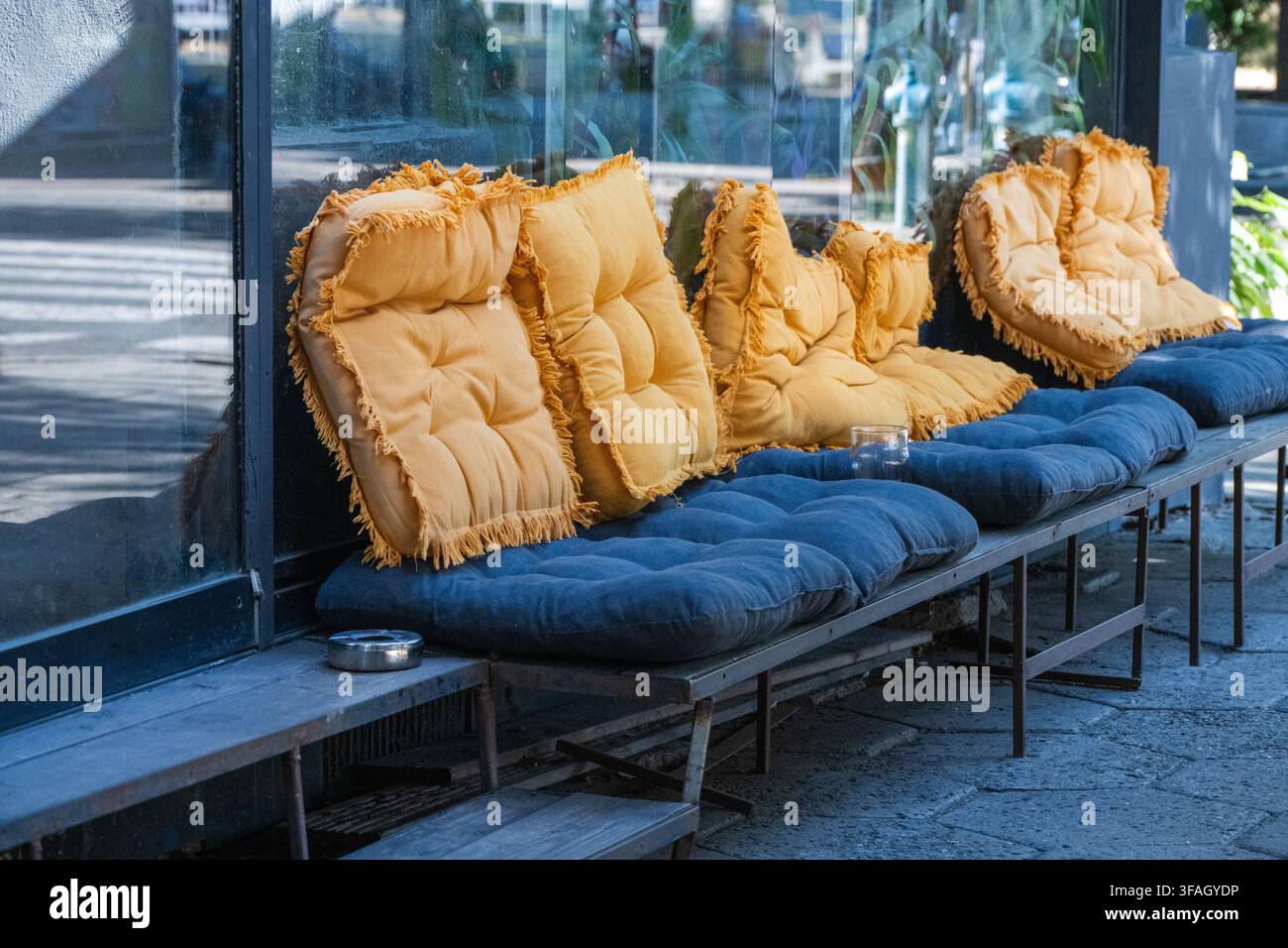 Une rangée de bancs de café en plein air présente des coussins d'assise bleus touffetés confortables et des oreillers de dossier jaunes moutarde à franges placés devant un verre réfléchissant Banque D'Images