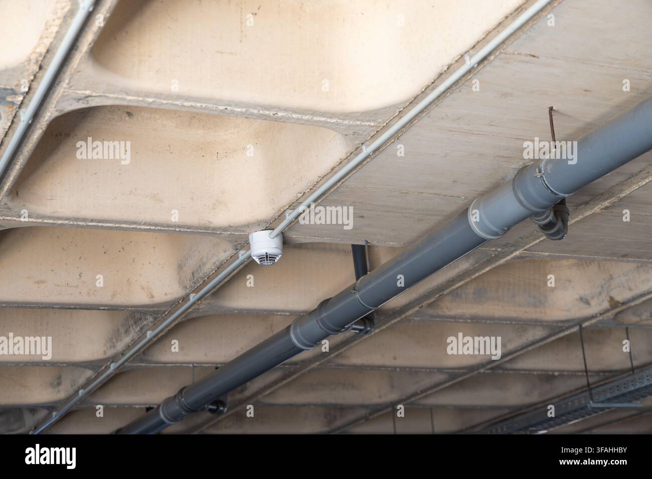 Une vue orientée vers le haut d'un plafond en béton avec des tuyaux visibles et un dispositif détecteur de fumée blanc fixé à la structure Banque D'Images