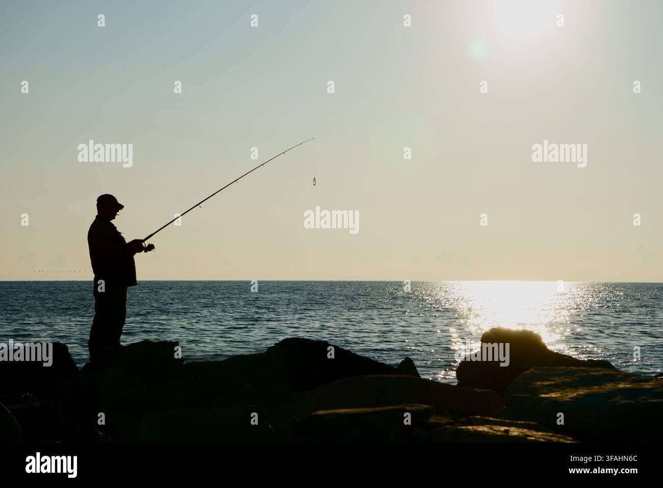 Silhouette d'un pêcheur solitaire debout sur une digue rocheuse tenant une canne à pêche contre une mer baignée de soleil. L'homme lance sa ligne dans Banque D'Images