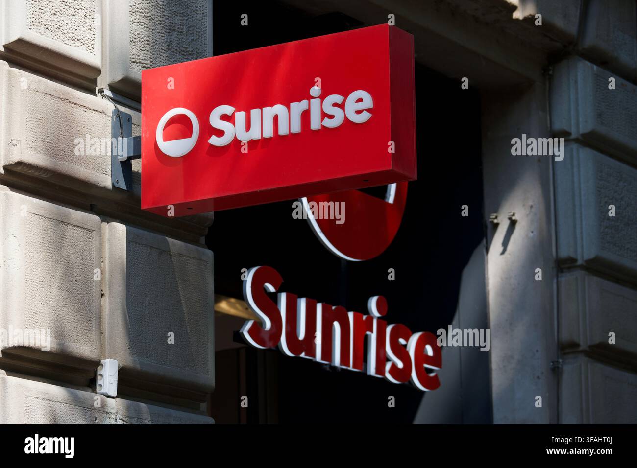 Zürich, Suisse - 30 juillet 2026 : Vue rapprochée en angle du panneau rouge du logo du magasin Sunrise accroché à la façade en pierre d’un bâtiment devant une boutique Banque D'Images
