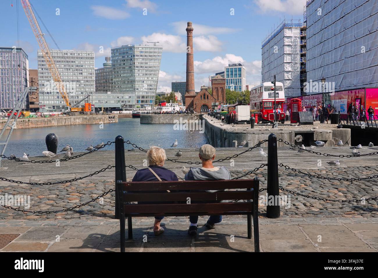 Vue arrière d'un couple de personnes âgées assis sur un banc au Royal Albert Dock à Liverpool, Royaume-Uni, regardant au loin sur l'eau vers le pub The Pump House Banque D'Images
